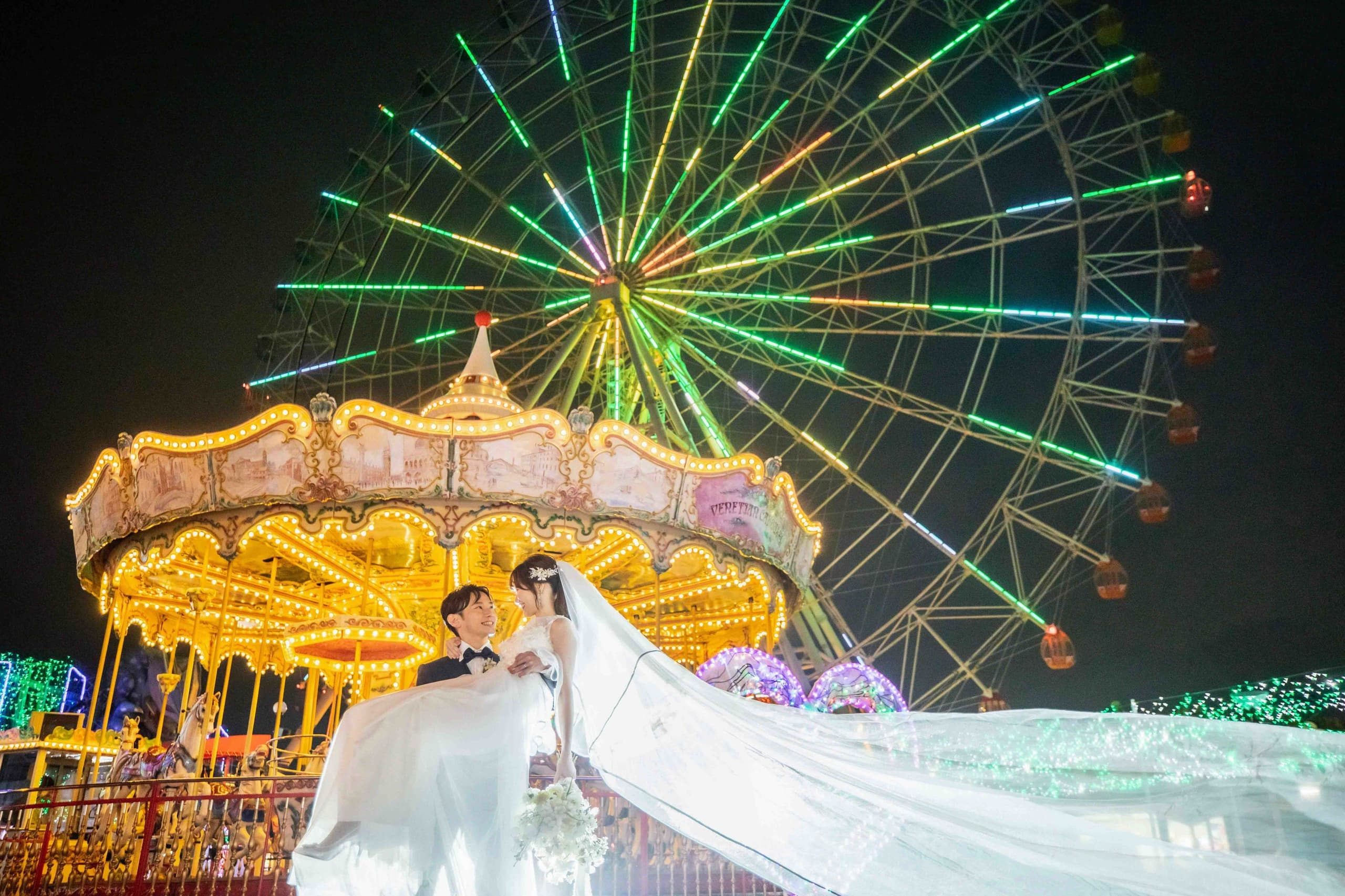 alt="夜の遊園地でナイトドレス前撮りをする笑顔の新郎新婦"